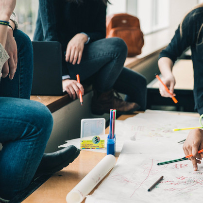 Brainstorming Vier Menschen sitzen an einem Tisch, die Gesichter sind nicht im Bild. Sie halten Stifte in der Hand und sehen nachdenkend aus. Eine Hand schreibt etwas auf das Plakat auf dem Tisch, welches sehr unstrukturiert gestaltet ist. Auf dem Tisch sind noch mehr Stifte und eine Tasse. Auf der Sitzbank erkennt man einen geöffneten Laptop und einen Rucksack im Hintergrund.