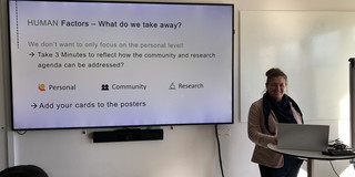 Frauke Mörike steht an einem kleinen Stehtisch mit Laptop und präsentiert in einem Seminarraum. Neben ihr hängt ein großer Bildschirm mit einer Präsentationsfolie mit dem Titel „HUMAN Factors – What do we take away?“. Auf der Folie steht unter anderem, dass nicht nur die persönliche Ebene betrachtet werden soll und dass drei Minuten Zeit zum Nachdenken über Community- und Forschungsaspekte genommen werden sollen. Außerdem sind die Punkte „Personal“, „Community“ und „Research“ aufgeführt sowie die Aufforderung, Karten zu den Postern hinzuzufügen.