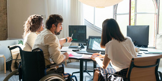 Drei Personen, bestehend aus einem Mann mit Rollstuhl und zwei Frauen, arbeiten kooperativ an mehreren Bildschirmen.