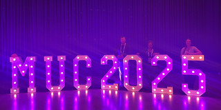 Violet illuminated letters stand out against a purple background. Behind them are Hendrik Sellmann, Frauke Mörike, and Jana-Sophie Effert leaning against the illuminated letters.